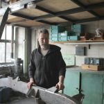 Bearded man working on metal piece in a rustic workshop setting.